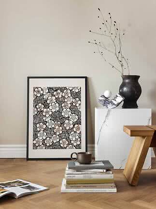 Lámina floral enmarcada apoyada en la pared junto a un jarrón, libros y taburete de madera