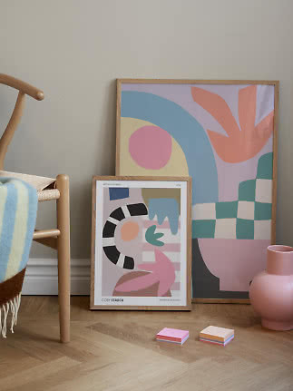 Dos láminas abstractas coloridas con marco de madera apoyadas en la pared junto a silla y jarrón rosa