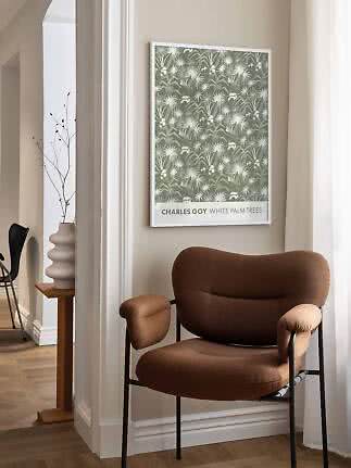 Affiche encadrée verte à palmiers blancs au-dessus d’un fauteuil brun dans une pièce lumineuse.