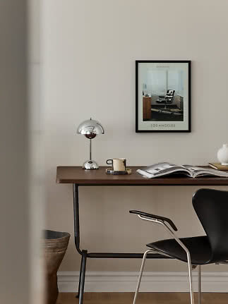 Minimalistisk skrivebord med kromlampe, kop, åben bog og sort stol ved beige væg med indrammet plakat