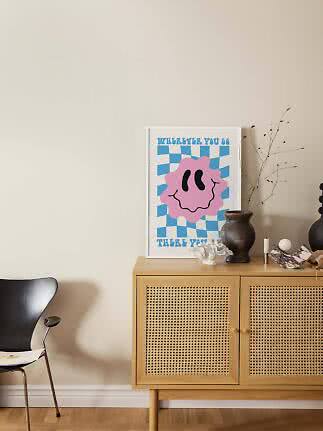 Colorful poster with pink smiley on blue checkered background, leaning on a wooden cabinet with vase.