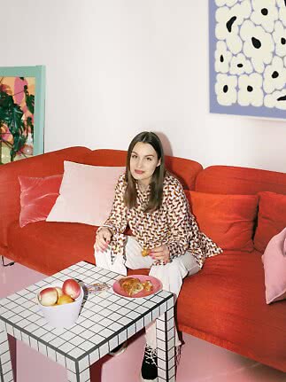 Vrouw zit op een rode bank met een croissant bij een geruite salontafel met fruit in kleurrijke kamer