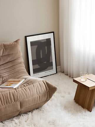 Poster astratto con cornice nera appoggiato al muro accanto a poltrona beige e tavolino in legno.