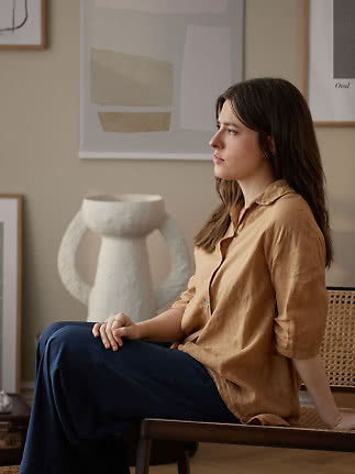 Kvinde i brun skjorte og mørke bukser sidder på bænk i stue med abstrakt kunst og hvid vase