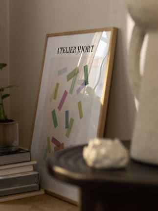 Poster Atelier Hjort incorniciato con forme colorate, appoggiato al muro accanto a libri e vaso