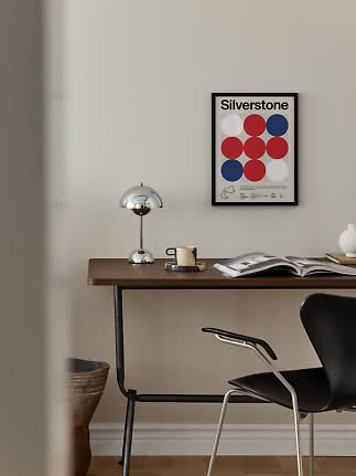 Dark wood desk with chrome lamp, mug and open book beneath a Silverstone poster on a light wall