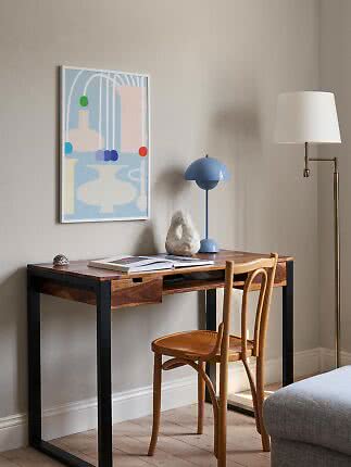 Wood desk with black frame, chair, blue table lamp and abstract wall art on a beige wall