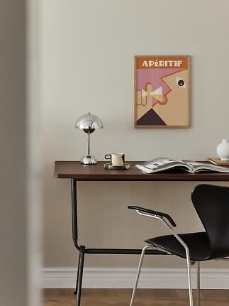 Minimalistischer Schreibtisch mit Chromlampe, Tasse, offenem Buch und gerahmtem Poster an beiger Wand.
