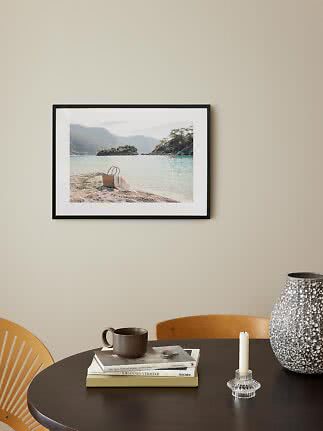 Cuadro con foto de playa y cesta junto al agua sobre mesa oscura con jarrón, vela y taza