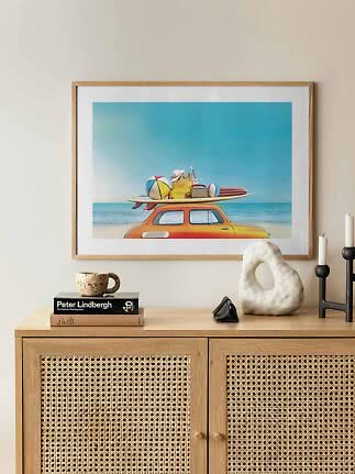 Framed beach print with orange car, surfboard, beach ball and suitcase under a blue sky