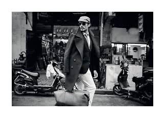 Black-and-white photo of stylish man in coat, cap and sunglasses carrying a large leather bag