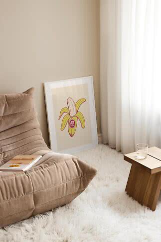Gerahmtes Poster mit gelber Bananenillustration, an beige Wand neben Sessel und Vorhang gelehnt