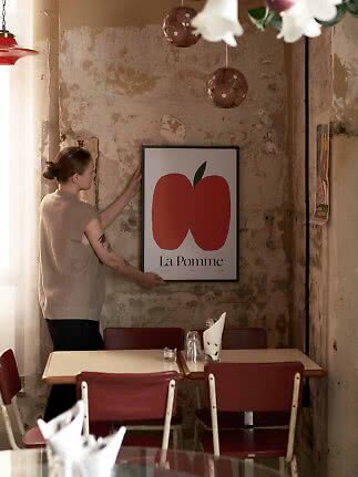 Person hängt La Pomme-Poster mit rotem Apfelmotiv an eine rustikale Caféwand.
