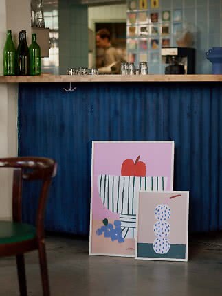 Two colorful art prints leaning against a blue café bar, with bottles and glasses in the background.