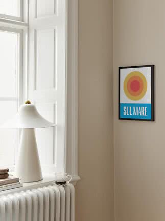 Framed SUL MARE poster with colored circles on a beige wall beside a window and white lamp