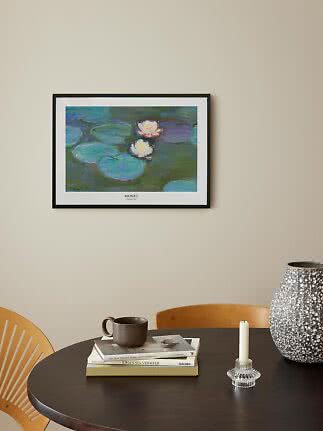 Gerahmter Monet-Seerosen-Druck über dunklem Tisch mit Büchern, Becher, Kerzenhalter und großer Vase.