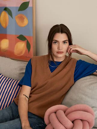 Kvinna i brun stickad väst över blå T-shirt sitter i en soffa bredvid en färgglad citrontavla.