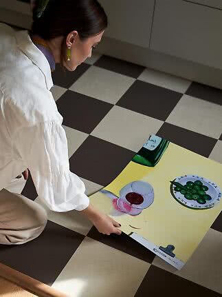 Woman on checkered floor holding a poster with a still life of green olives, glass and bottle.