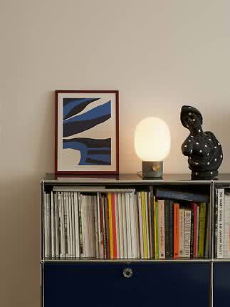 Lit table lamp on shelf beside framed art print, black sculpture and books
