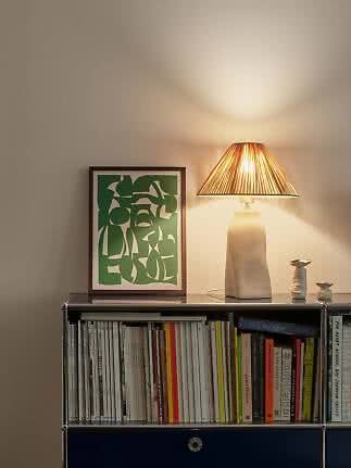 Lampada da tavolo con paralume plissettato su libreria accanto a stampa verde incorniciata e piccoli vasi