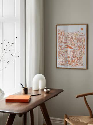 Framed art print with orange landscape illustration above a wooden desk by a window
