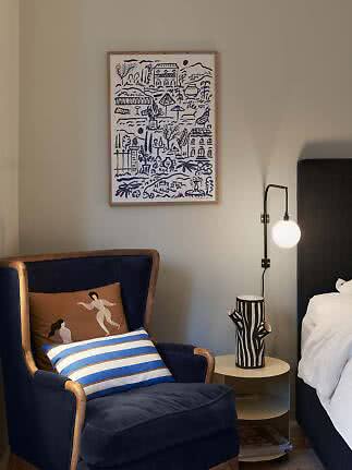 Framed blue and white illustration above a navy armchair beside a side table and lamp in a bedroom