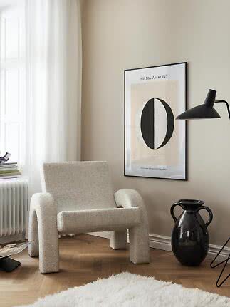 Hyggeligt hjørne med cremefarvet lænestol, sort væglampe, indrammet kunst og sort vase
