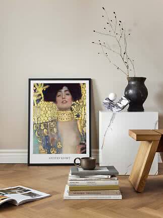 Inramad Gustav Klimt-affisch mot väggen med böcker, mugg och svart vas på vit piedestal