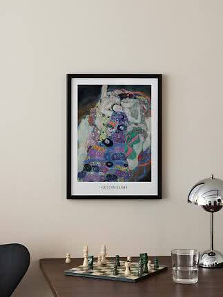 Inramad Gustav Klimt-affisch ovanför ett bord med schackbräde, vattenglas och kromlampa