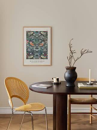 Inramad William Morris-affisch med fåglar och blommor ovanför ett mörkt matbord med vas och ljus.