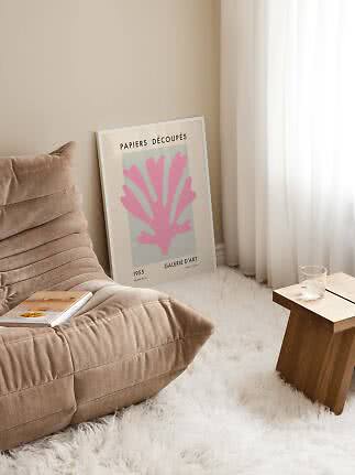 Framed art print with pink motif leaning by the wall beside a beige chair and small wooden table