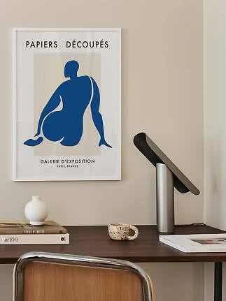 Framed art print with blue abstract figure above a desk with lamp, books, vase and mug