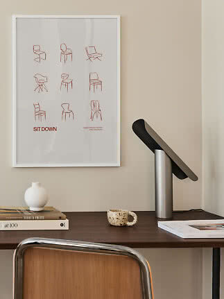 Minimalistisk skrivebord med sort bordlampe, bøger, vase og indrammet stolplakat på væggen