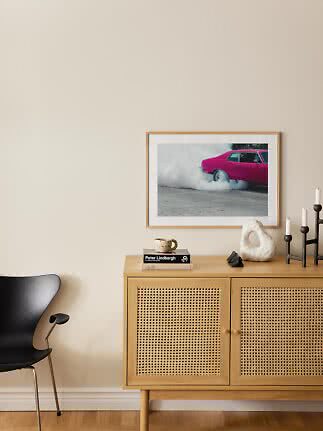 Light wood sideboard with cane doors, black chair and framed photo of a pink car on a beige wall
