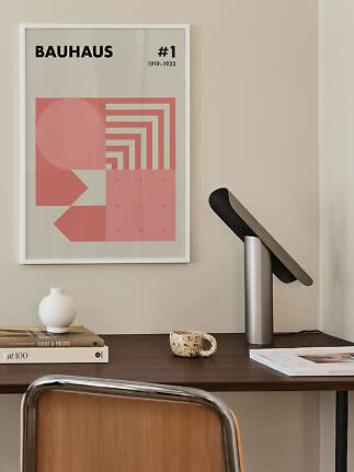 Skrivebord med sort bordlampe og indrammet Bauhaus-plakat i rosa toner på væggen