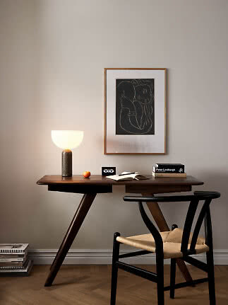 Scrivania in legno scuro con lampada accesa, libri, taccuino e sedia nera sotto un quadro.