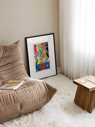Farverig indrammet kunstplakat læner sig op ad væggen ved lænestol, gardiner og lille træbord