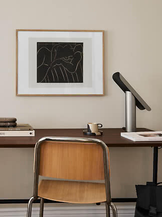 Minimalistisk hjemmekontor med træstol, skrivebord, lampe og indrammet abstrakt kunst på væggen.