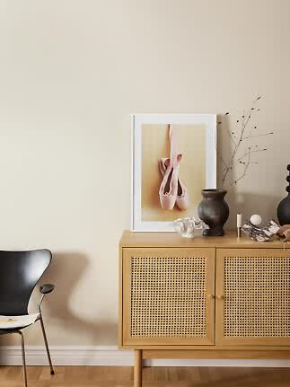 Light wooden cabinet with cane doors, vase, and framed photo of pink ballet shoes against beige wall