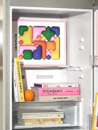 Offener Kühlschrank mit buntem abstraktem Kunstwerk, Bücherstapeln auf Fächern und einer Orange.