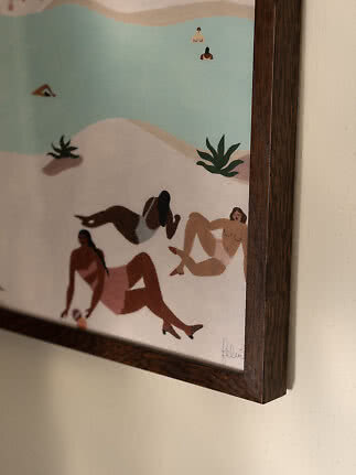 Framed illustration of women relaxing by a light blue lake on a sandy beach