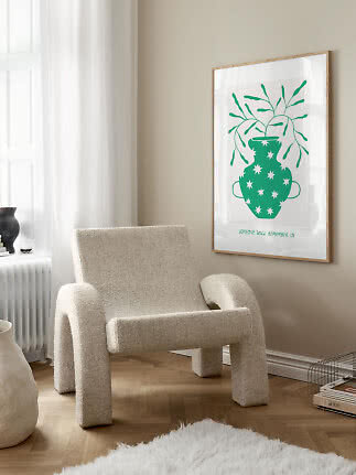 Framed poster with green vase and branches on beige wall beside a cream armchair
