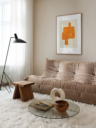 Beige sofa in living room with glass table, black floor lamp and abstract orange wall art