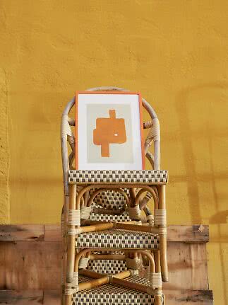 Framed abstract orange artwork on a woven rattan chair against a yellow wall