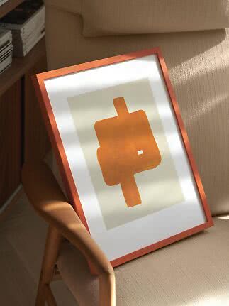 Framed abstract orange art print leaning on a beige armchair in warm sunlight