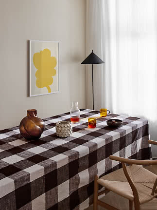 Spisebord med brun- og hvidternet dug, glasvaser, kopper og sort gulvlampe ved vindue.