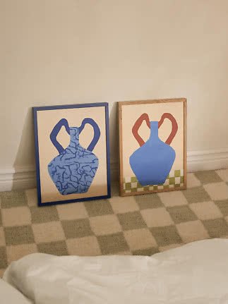 Deux affiches encadrées de vases bleus appuyées contre un mur beige sur un tapis à carreaux.