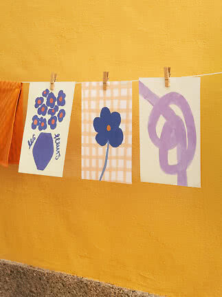 Drei Kunstdrucke hängen an einer Leine vor gelber Wand, mit violetten Blumen und abstrakten Formen.