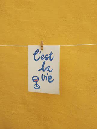 White card with blue "C'est la vie" text and wine glass, clipped to a line against a yellow wall