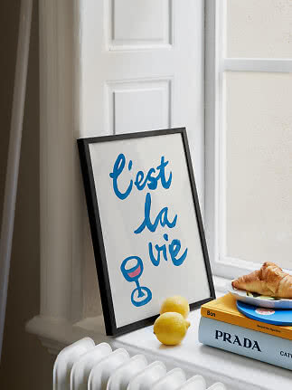 Framed "c'est la vie" print on a windowsill with lemons, books and a croissant.